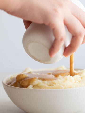 Foto de perfil de una mano echando salsa gravy vegana sobre un bol de puré de patatas