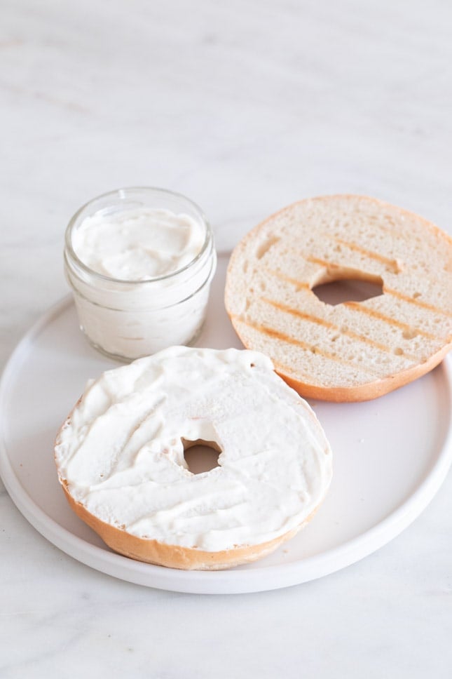 Foto de un plato con un tarro con queso crema vegano y un par de rebanadas de bagel untadas con queso