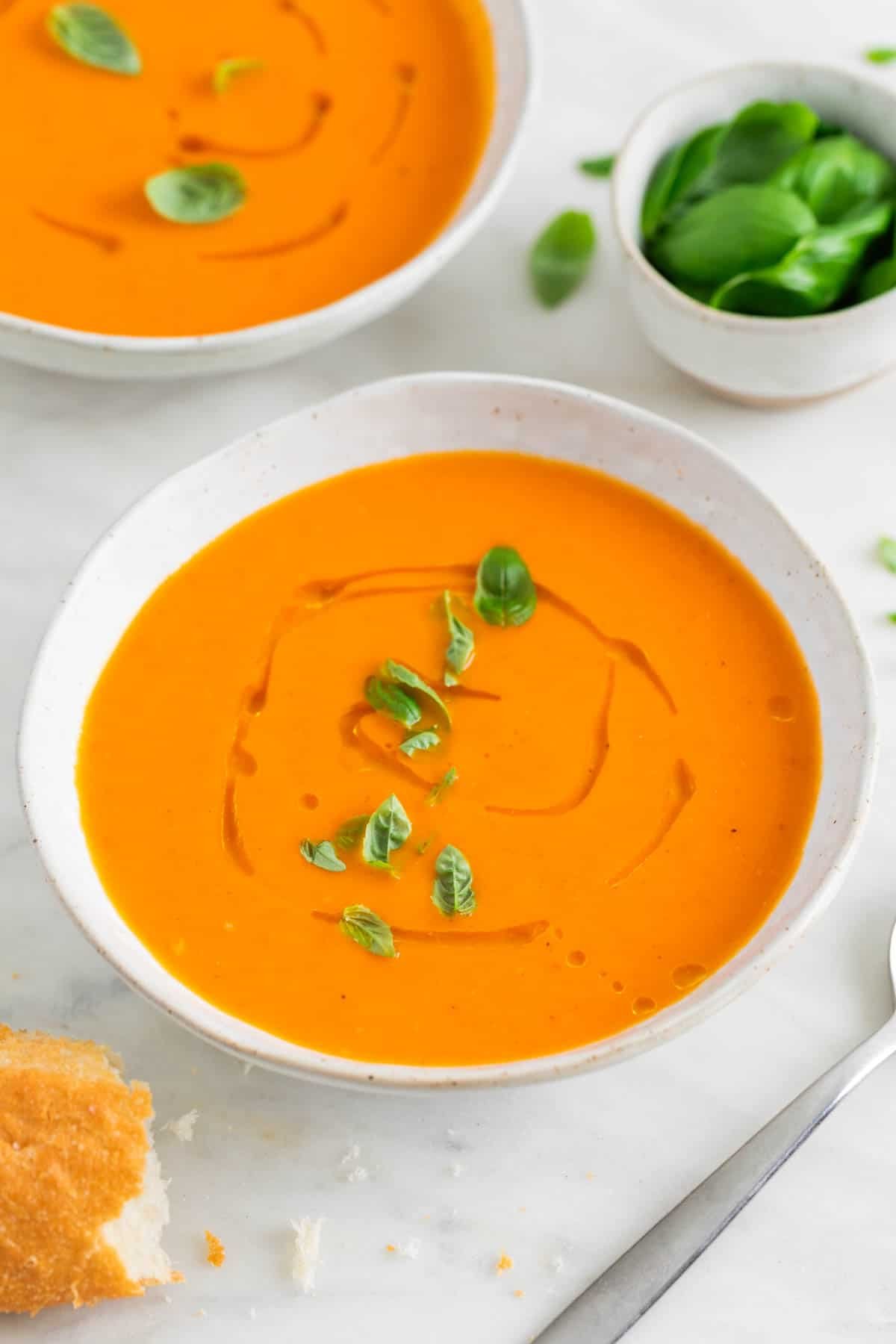 Dos boles con sopa de tomate decorados y un bol con albahaca y pan al fondo.