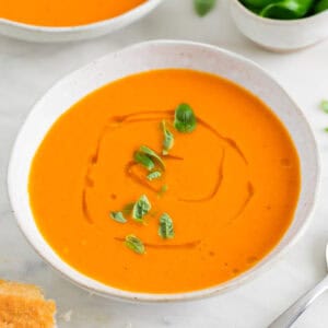 Dos boles con sopa de tomate decorados y un bol con albahaca y pan al fondo.
