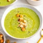 Bol de crema de calabacín decorado con una cuchara e ingredientes alrededor.