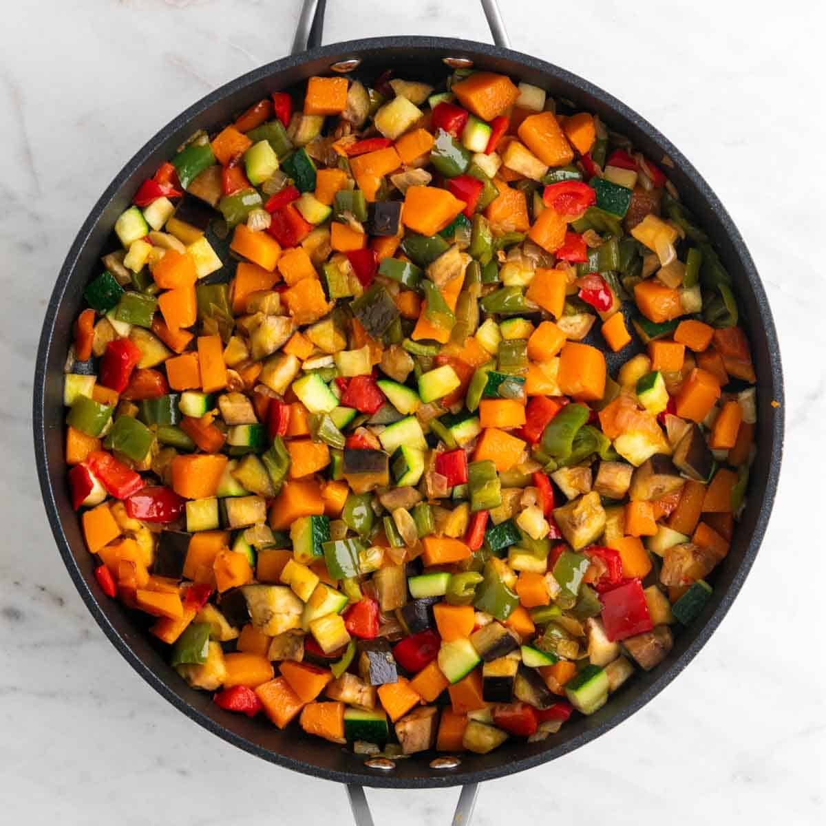 Sartén con cebolla, ajo, pimientos, calabaza, berenjena y calabacín cocinados.