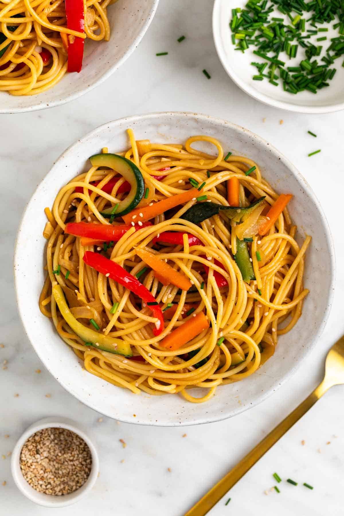 Dos platos de espaguetis con verduras, decorados con sésamo y cebollino, con un tenedor e ingredientes alrededor.