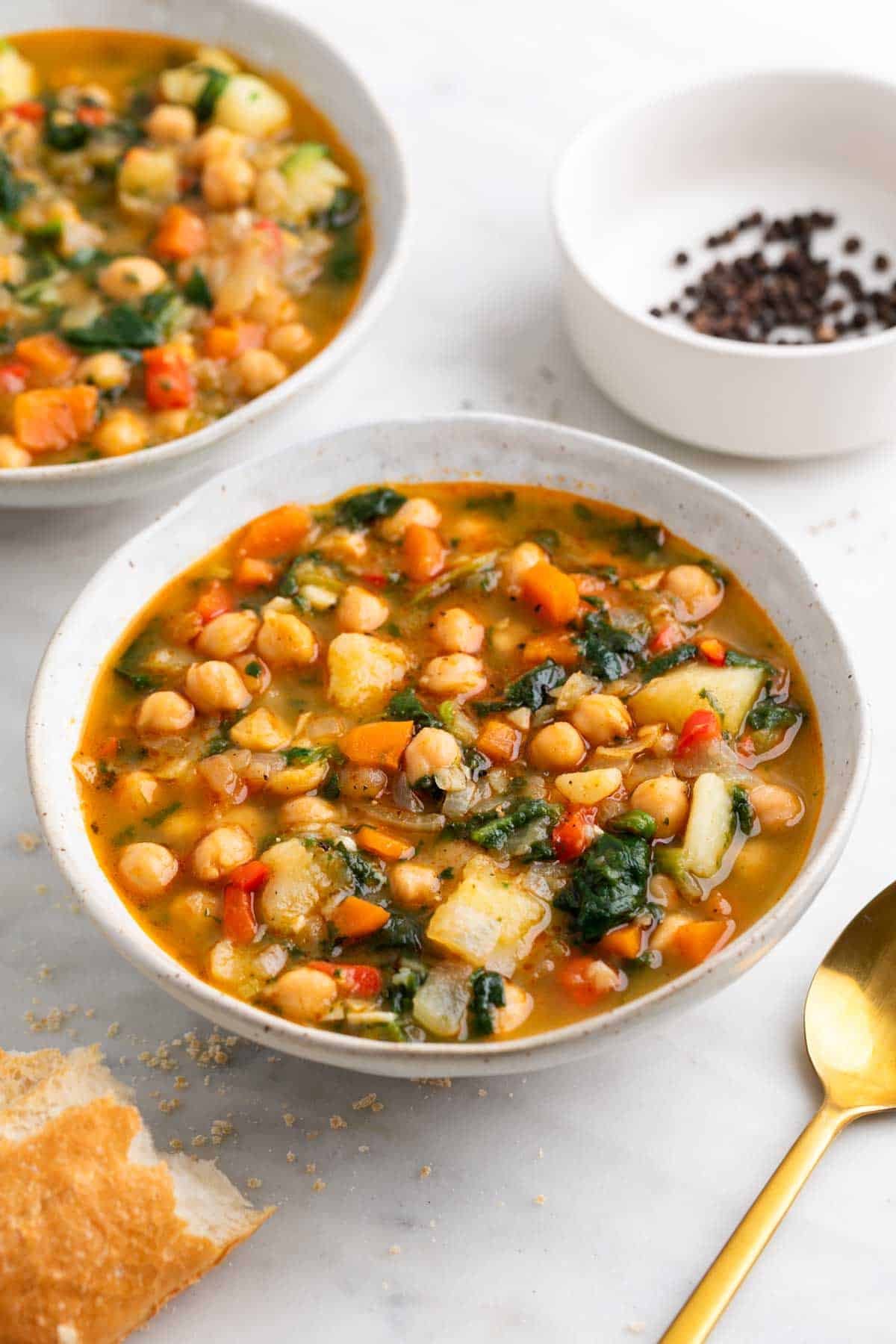 Cuencos de potaje de garbanzos con verduras acompañados de pan y una cuchara.