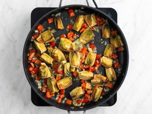 Cebolla, pimiento rojo, ajo y alcachofas salteados en una sartén.