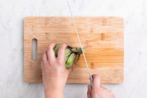 Manos troceando el extremo del tallo de una alcachofa con un cuchillo sobre una tabla.
