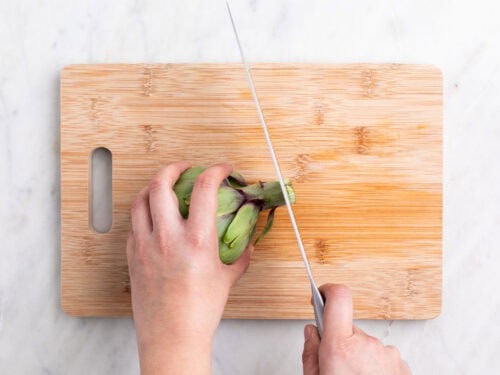 Manos troceando el extremo del tallo de una alcachofa con un cuchillo sobre una tabla.