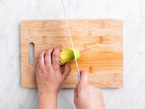 Manos troceado la punta de una alcachofa sin hojas con un cuchillo sobre una tabla.