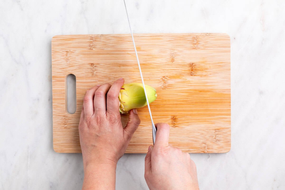 Manos troceado la punta de una alcachofa sin hojas con un cuchillo sobre una tabla.