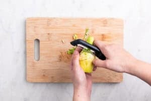 Manos pelando el tallo de una alcachofa con un pelador sobre una tabla.