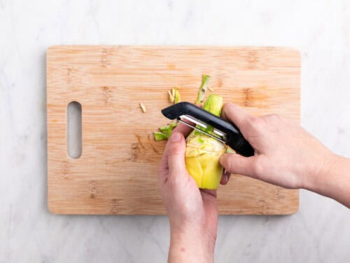 Manos pelando el tallo de una alcachofa con un pelador sobre una tabla.