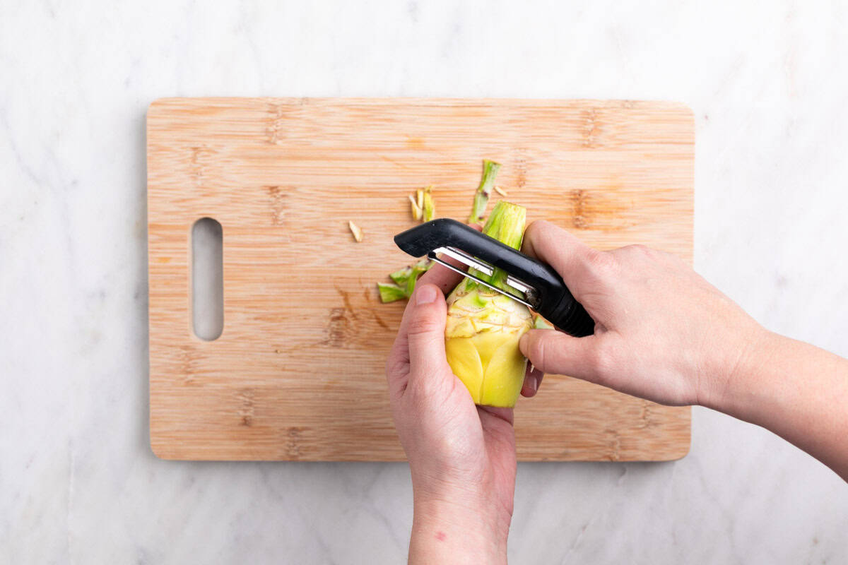 Manos pelando el tallo de una alcachofa con un pelador sobre una tabla.