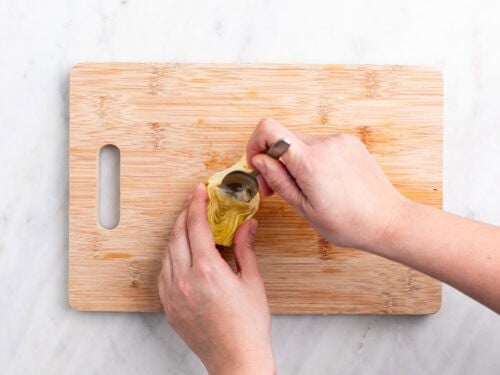 Mano limpiando los pelillos de una alcachofa con una cuchara sobre una tabla.