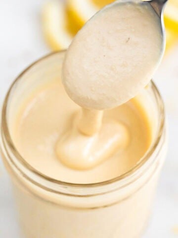 Aliño de tahini casero con textura cremosa en un bote, con una cuchara y rodajas de limón